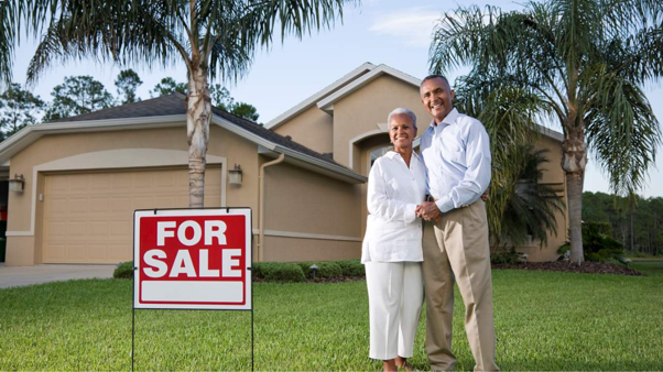 Older couple standing outside a house with a sold sign, considering a downsizer super contribution.